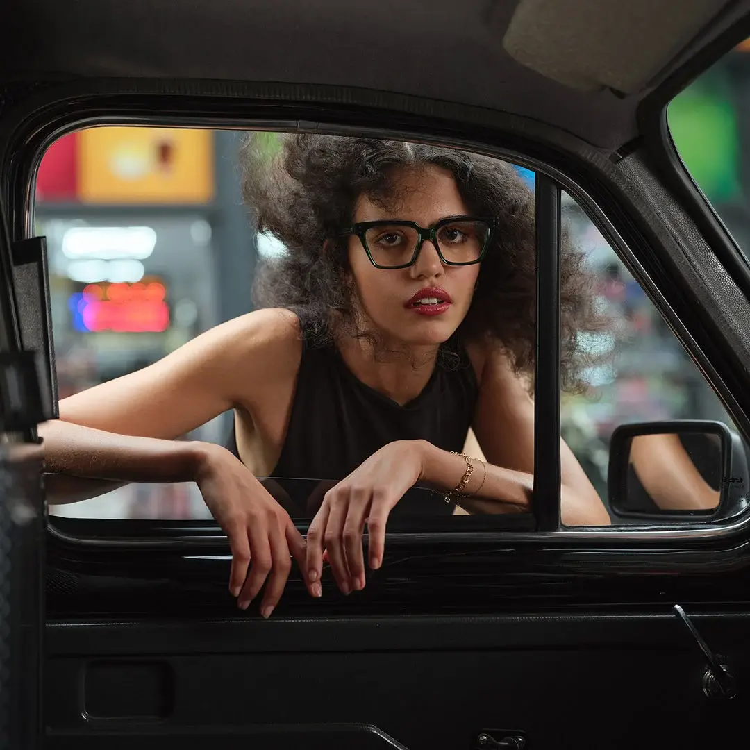 Femme en lunettes de vue noires Cutler and Gross, accoudée à la fenêtre d’une voiture la nuit